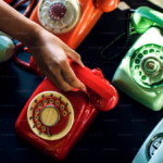 Hand reaching toward a red phone among a collection of dial and push-button phones on a table