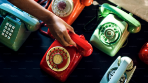 Hand reaching toward a red phone among a collection of dial and push-button phones on a table