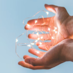 Two hands holding glowing fairy lights against a clear blue sky, symbolizing securing an organization’s future through planned giving.