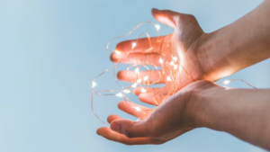 Two hands holding glowing fairy lights against a clear blue sky, symbolizing securing an organization’s future through planned giving.